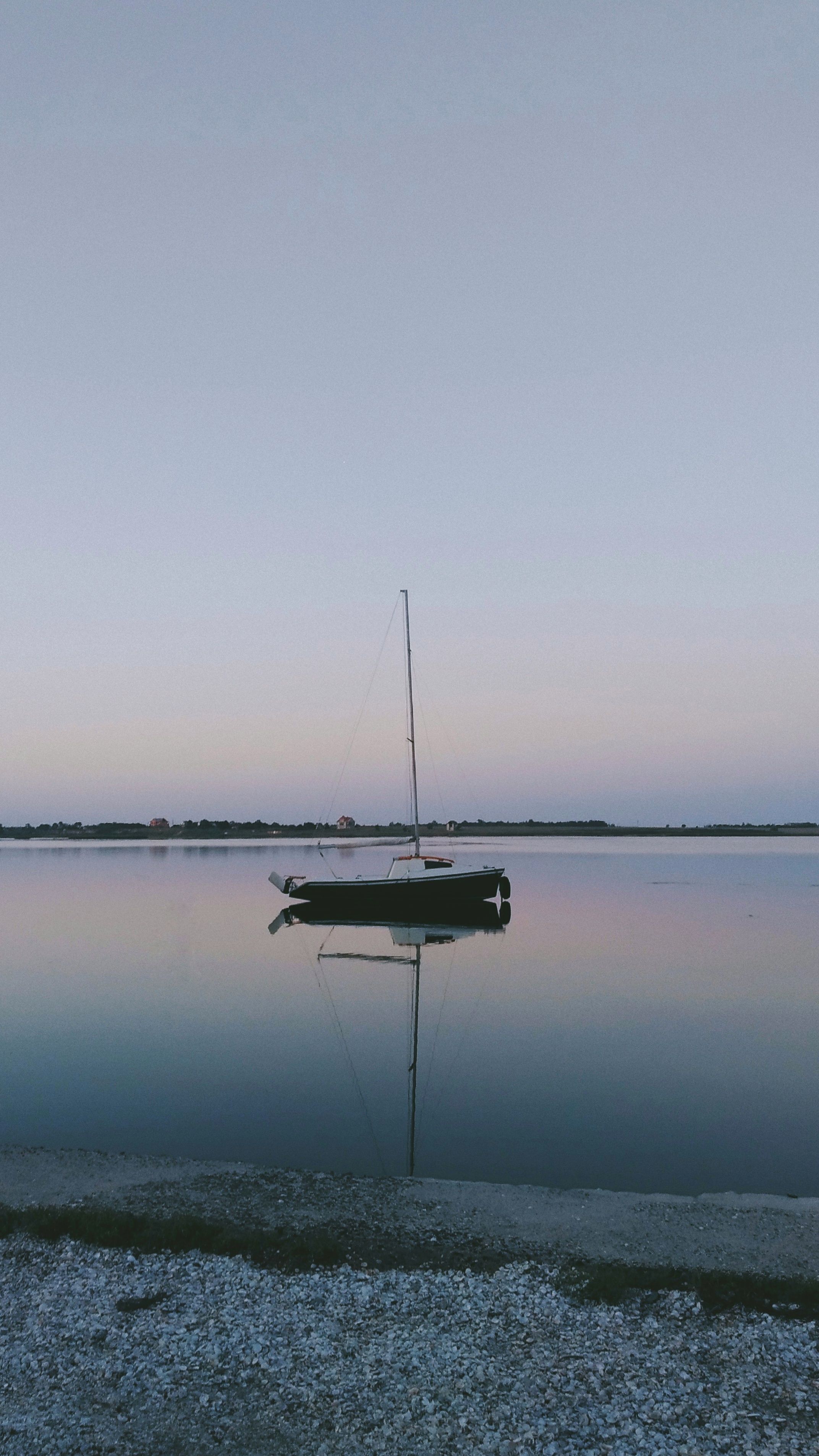 Tranquil scene of a sailboat and its reflection in calm waters at twilight, near a peaceful shoreline.