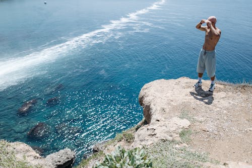 Free Shirtless Man in Gray Shorts Standing on Cliff Near Body of Water Stock Photo