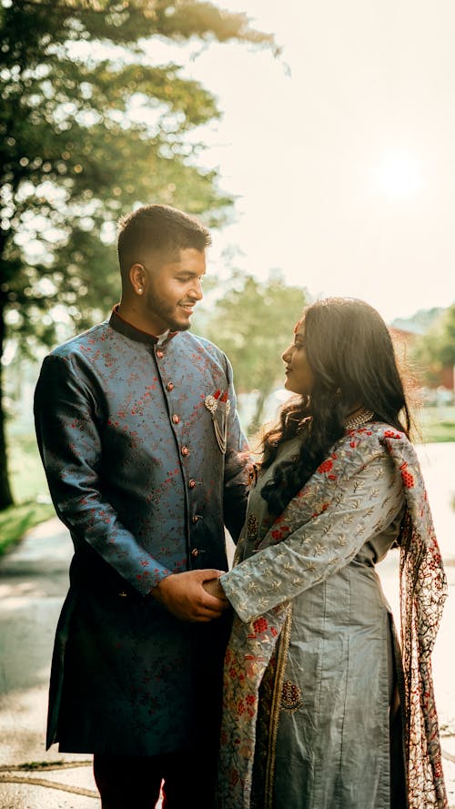 Side view of romantic Indian couple wearing traditional outfits holding hands while standing on street with trees during wedding celebration