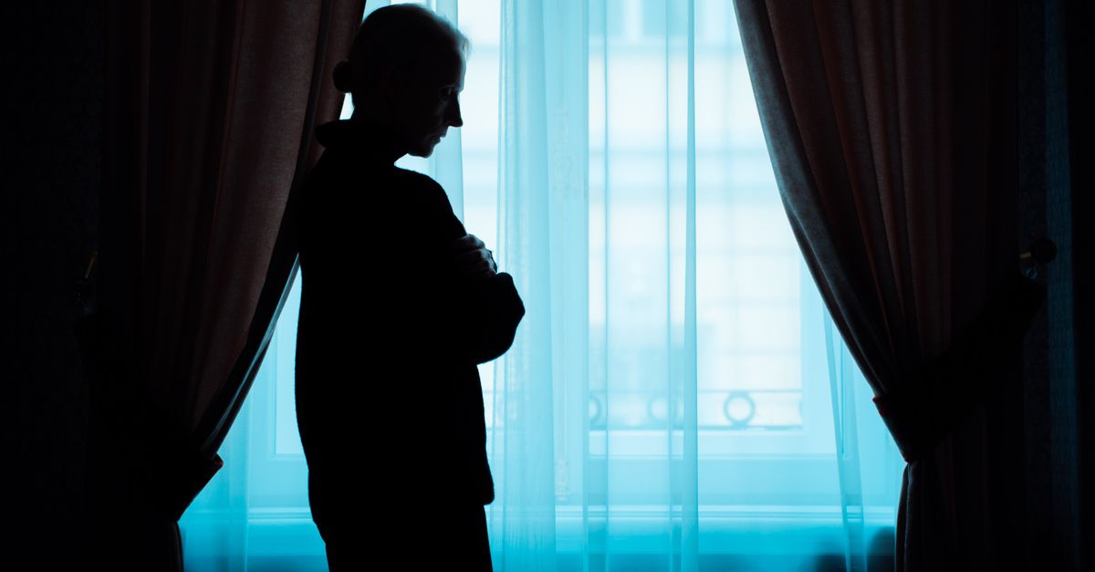A silhouette of a senior woman stands thoughtfully by a window, conveying solitude.