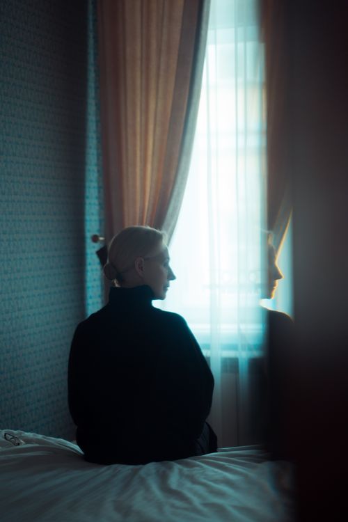Free Woman sits on bed, looking out window, in dimly lit room, reflecting solitude. Stock Photo