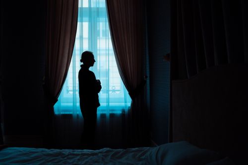 Free Moody silhouette of a woman standing alone by a window with curtains in a dimly lit room. Stock Photo