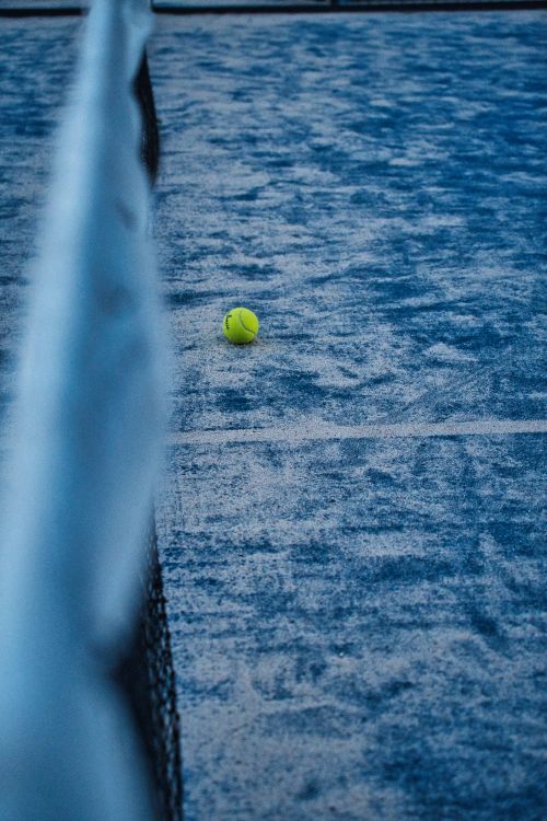 Free A lone tennis ball rests on a textured blue clay court at dusk, creating a serene athletic imagery. Stock Photo