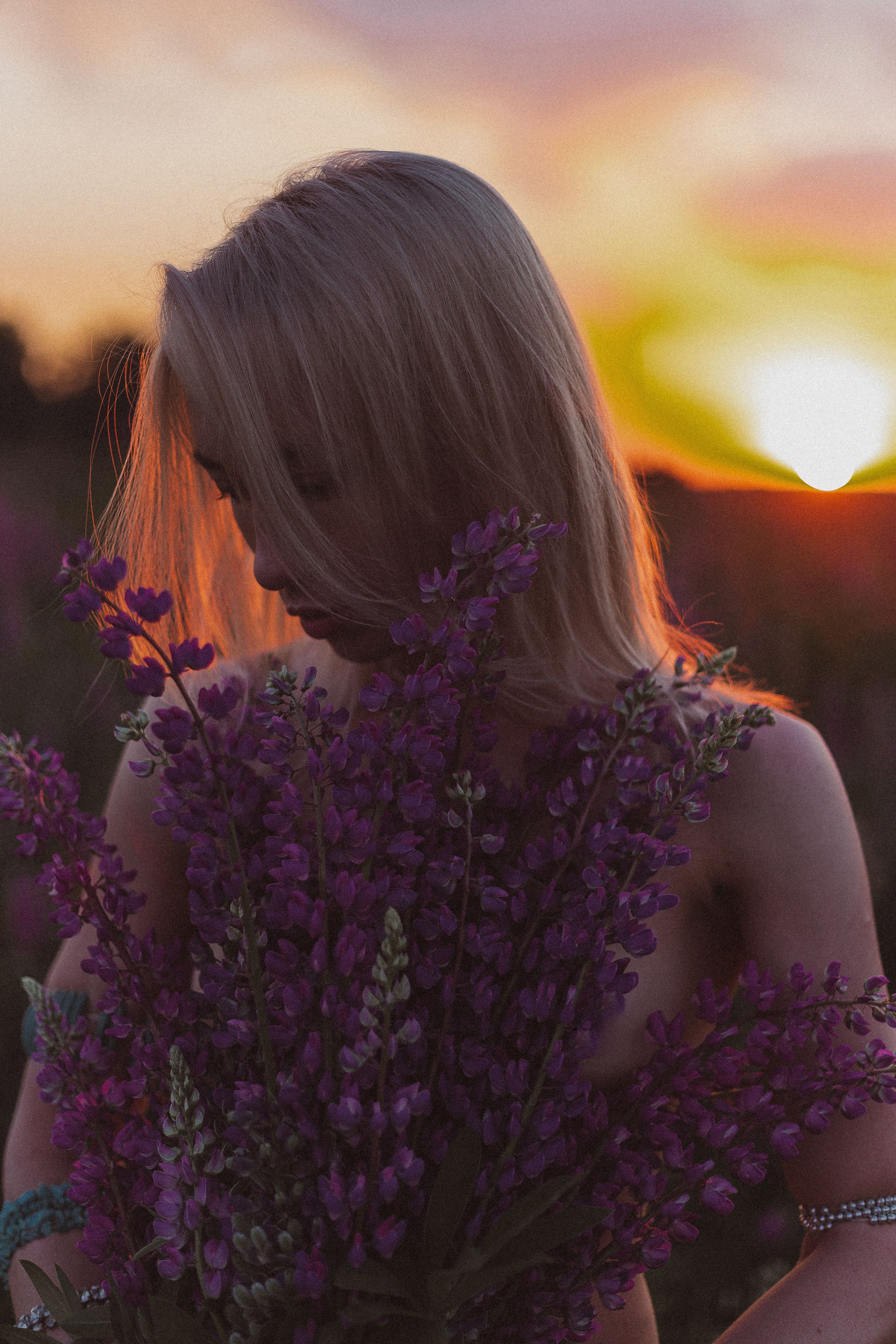 Blonde woman holding flowers, gazing down during a vibrant sunset.
