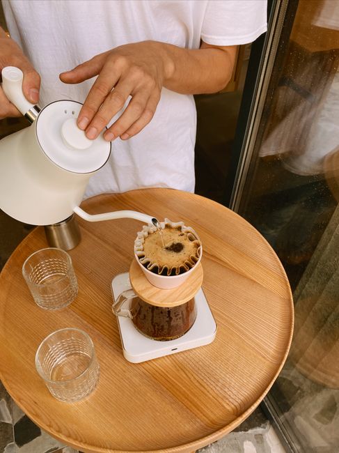 Hands brewing pour-over coffee with a kettle and dripper on a wooden table.