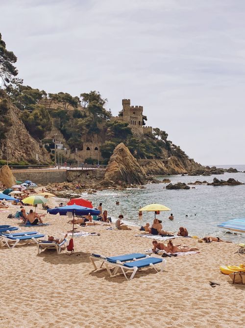 Free Vibrant beach scene in Lloret de Mar, Spain, featuring sunbathers and a historic castle. Stock Photo
