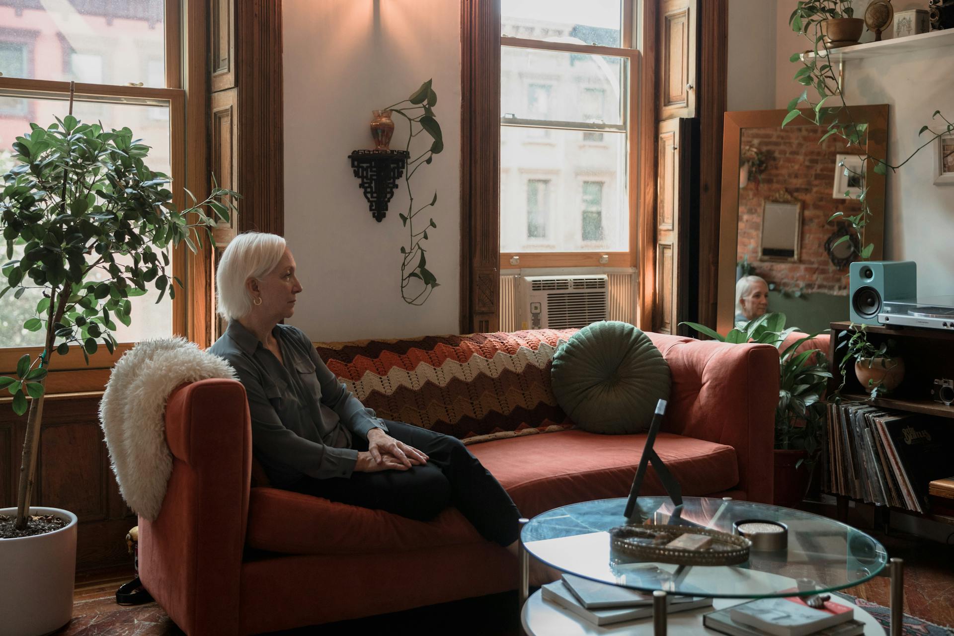 A senior woman with white hair sitting thoughtfully on a colorful sofa in a cozy living room.