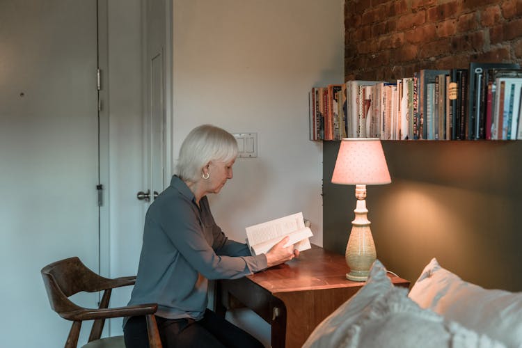 An Elderly Woman Reading A Book Near A Lamp