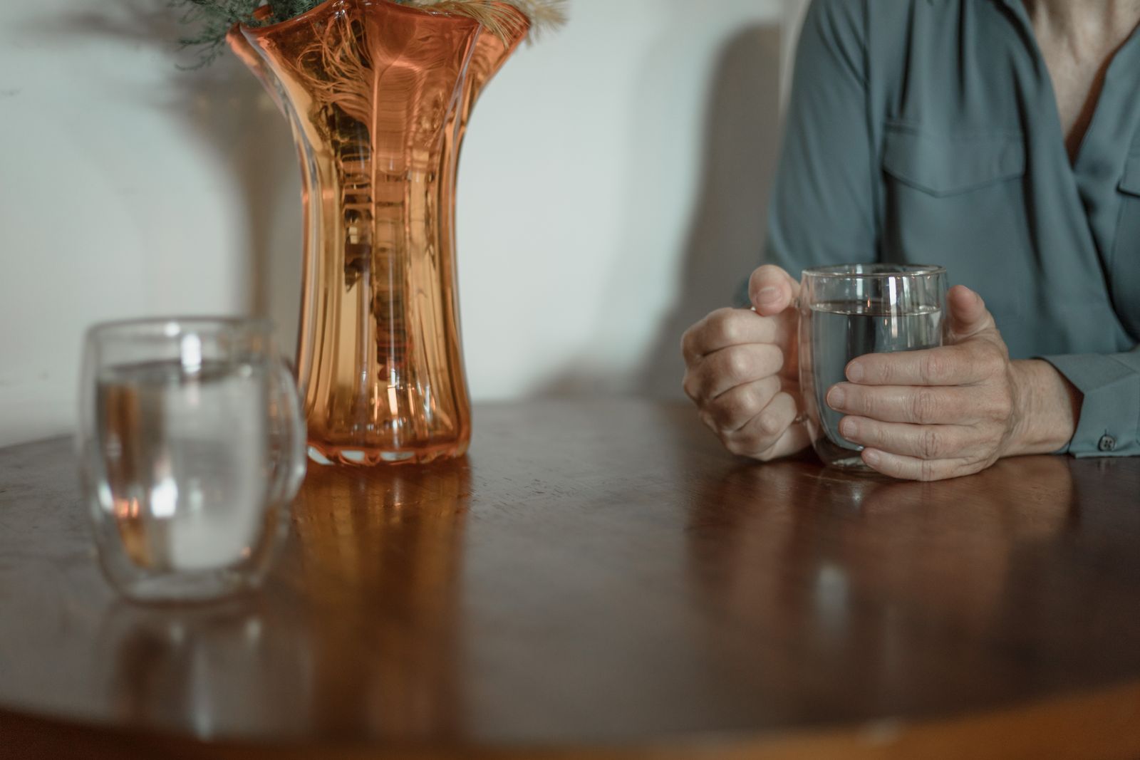 Hands holding a glass of water at a wooden table beside an amber glass vase and second glass.