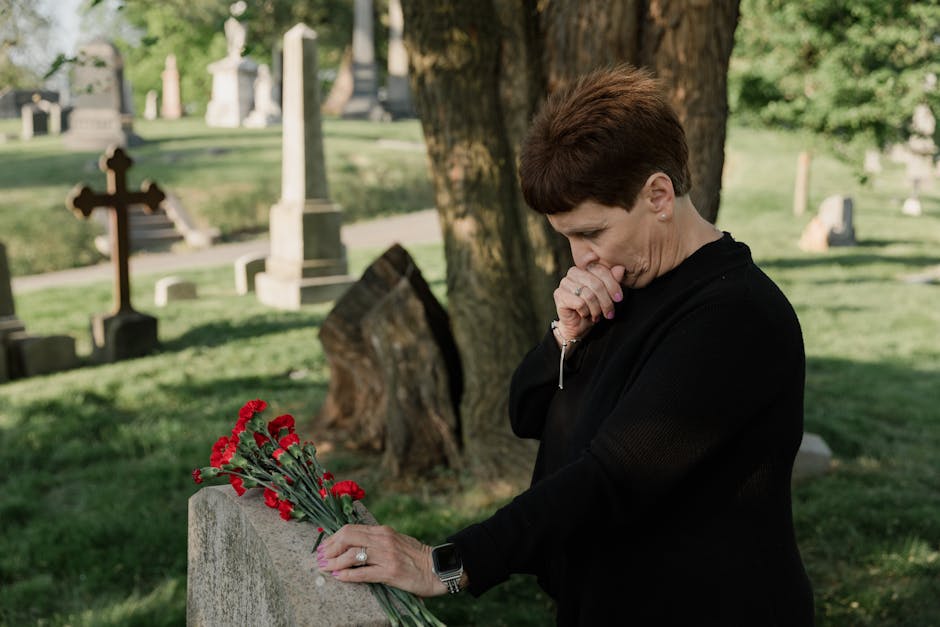 Senior woman in black at a cemetery, grieving by a gravestone with red flowers.
