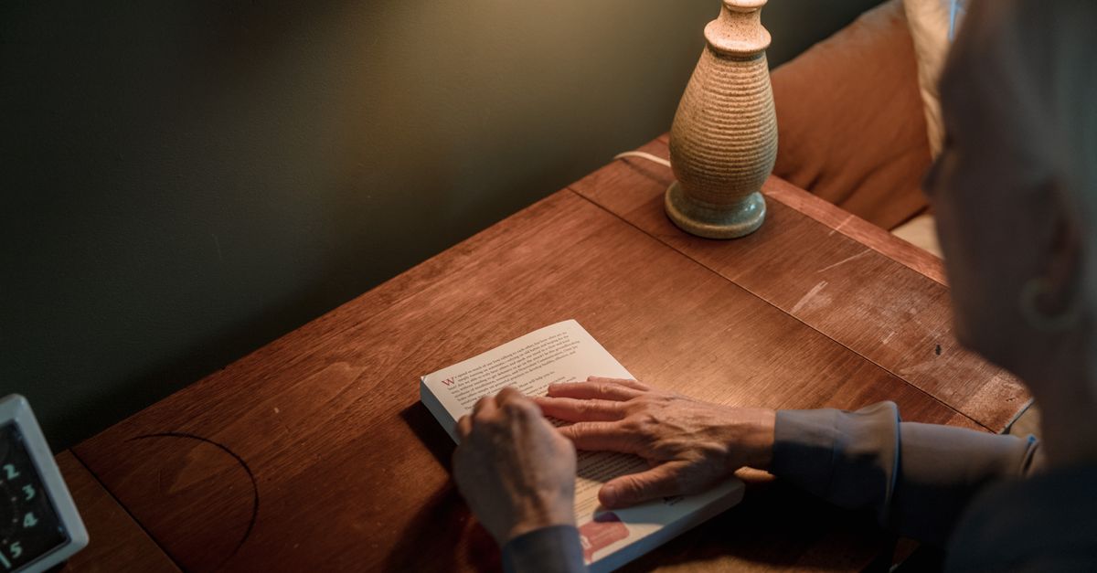 A senior woman reads a book on a wooden desk under warm lamp lighting.