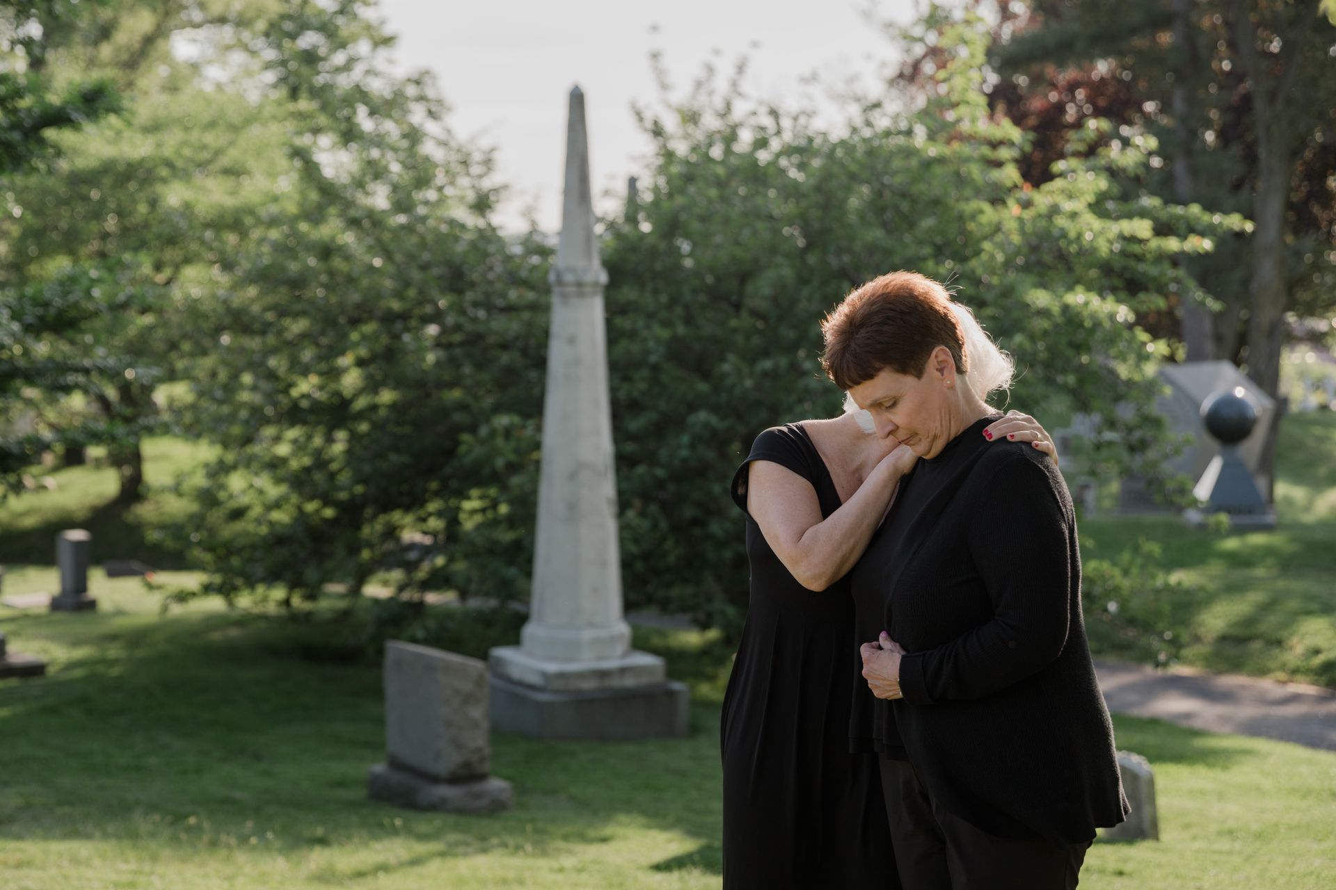 Two women in black embrace in a cemetery, expressing grief and support amidst serene greenery.
