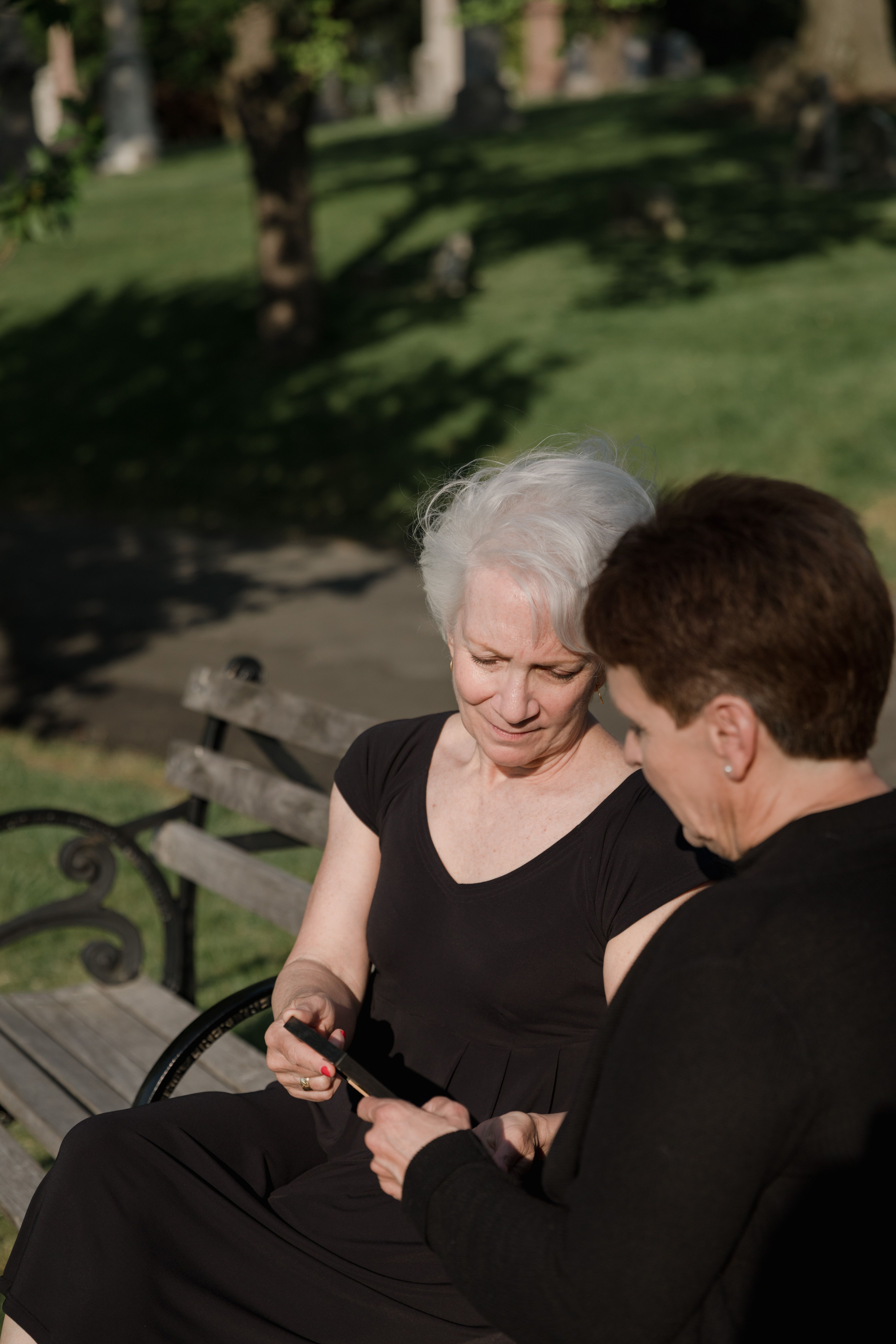 Two senior adults sitting on a park bench sharing a smartphone on a sunny day.
