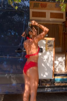 A woman in a red swimsuit enjoys an outdoor shower by a campervan under the sun.