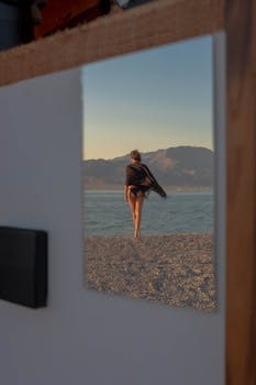 A woman in a scarf and bikini is reflected in a mirror, capturing a scenic beach view with mountains.