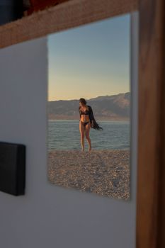 A woman in a bikini is reflected in a mirror on a beach at sunset, creating a serene summer scene.