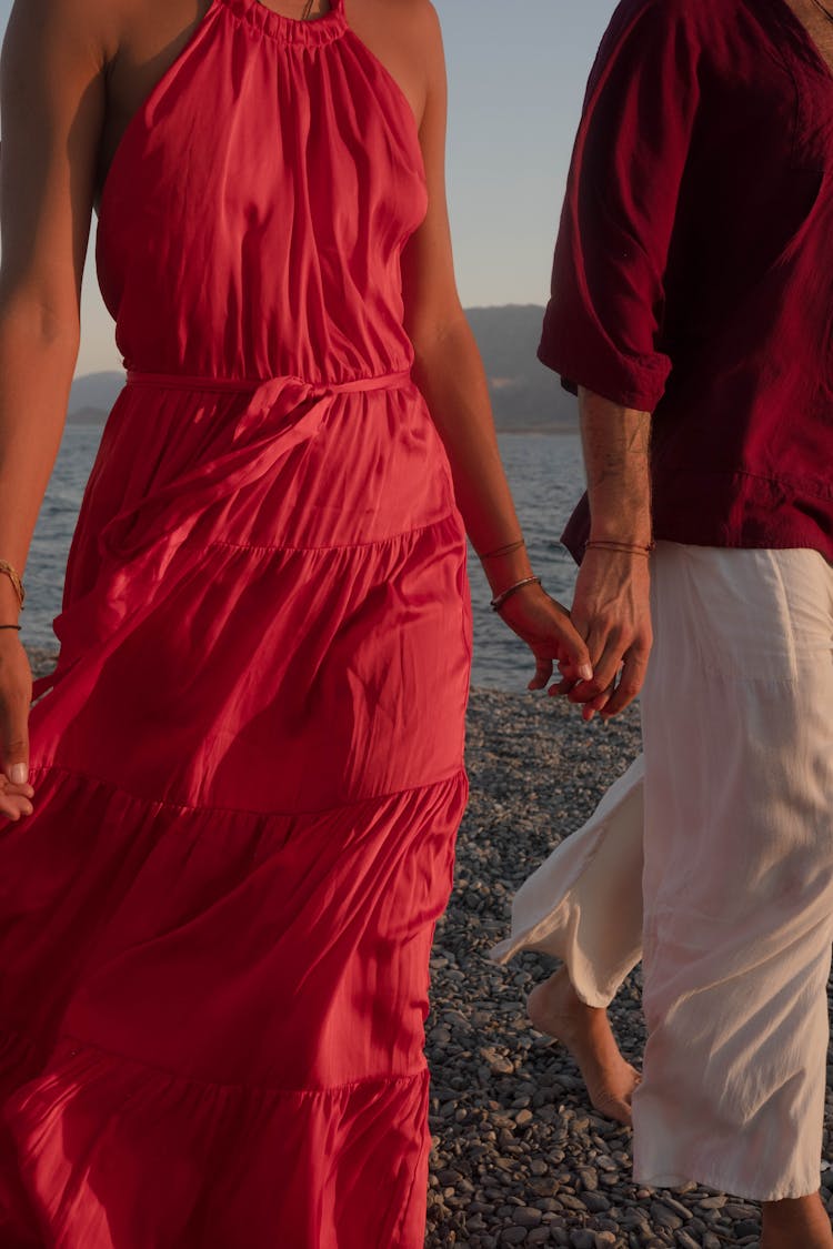 Man And Woman In Red Dress Walking Hand In Hand In The Beach