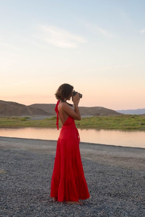 Free A woman in a red dress taking photos by a tranquil river at sunset. Stock Photo
