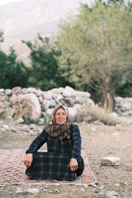 An elderly woman in a headscarf sitting peacefully on a rug outdoors, surrounded by nature.