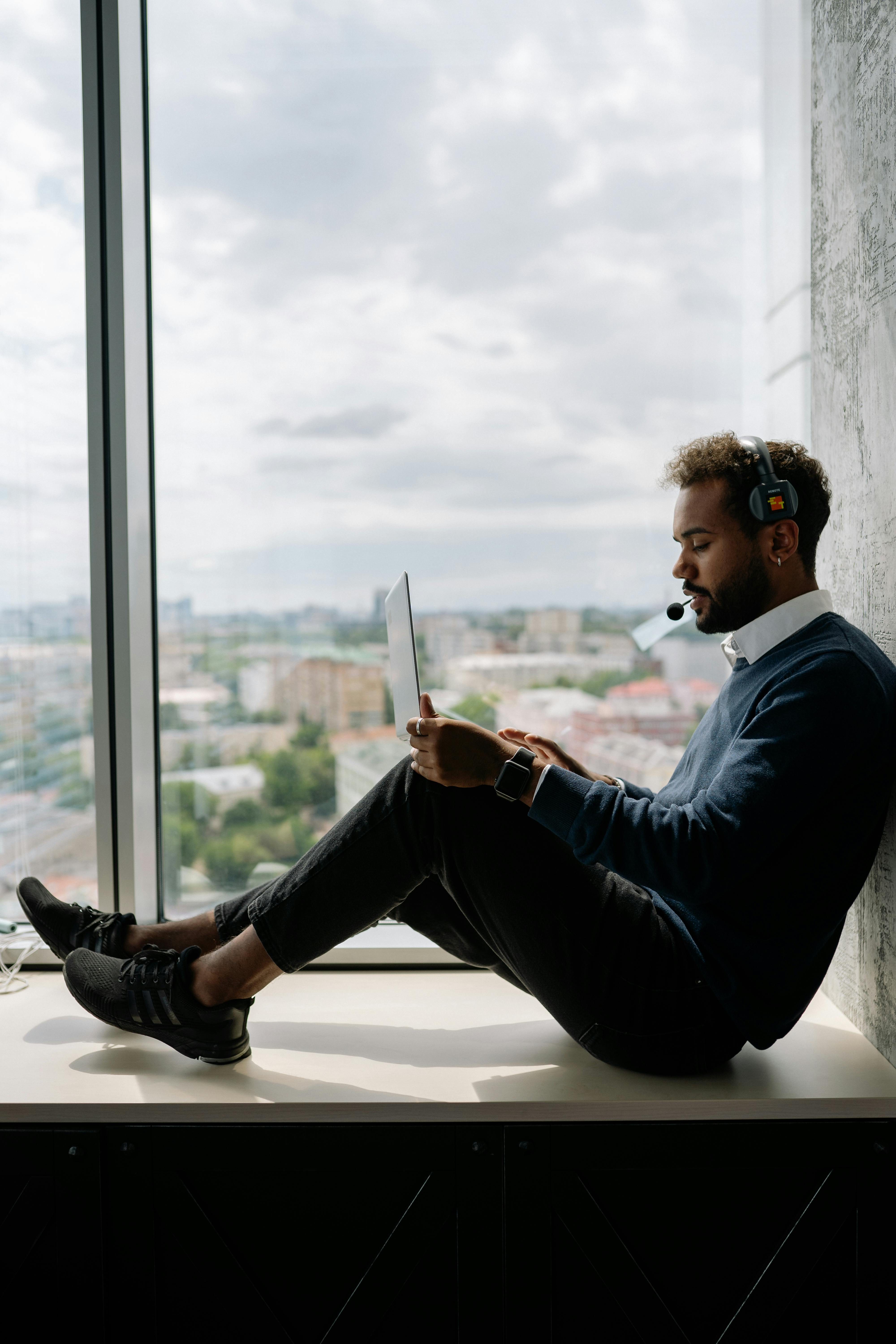 man using his laptop while sitting on the window