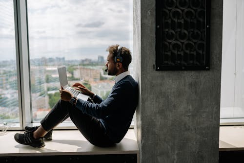 Man Using His Laptop while Sitting on the Window