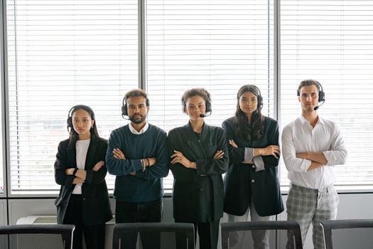 A diverse group of call center agents standing with arms crossed, wearing headsets in an office.