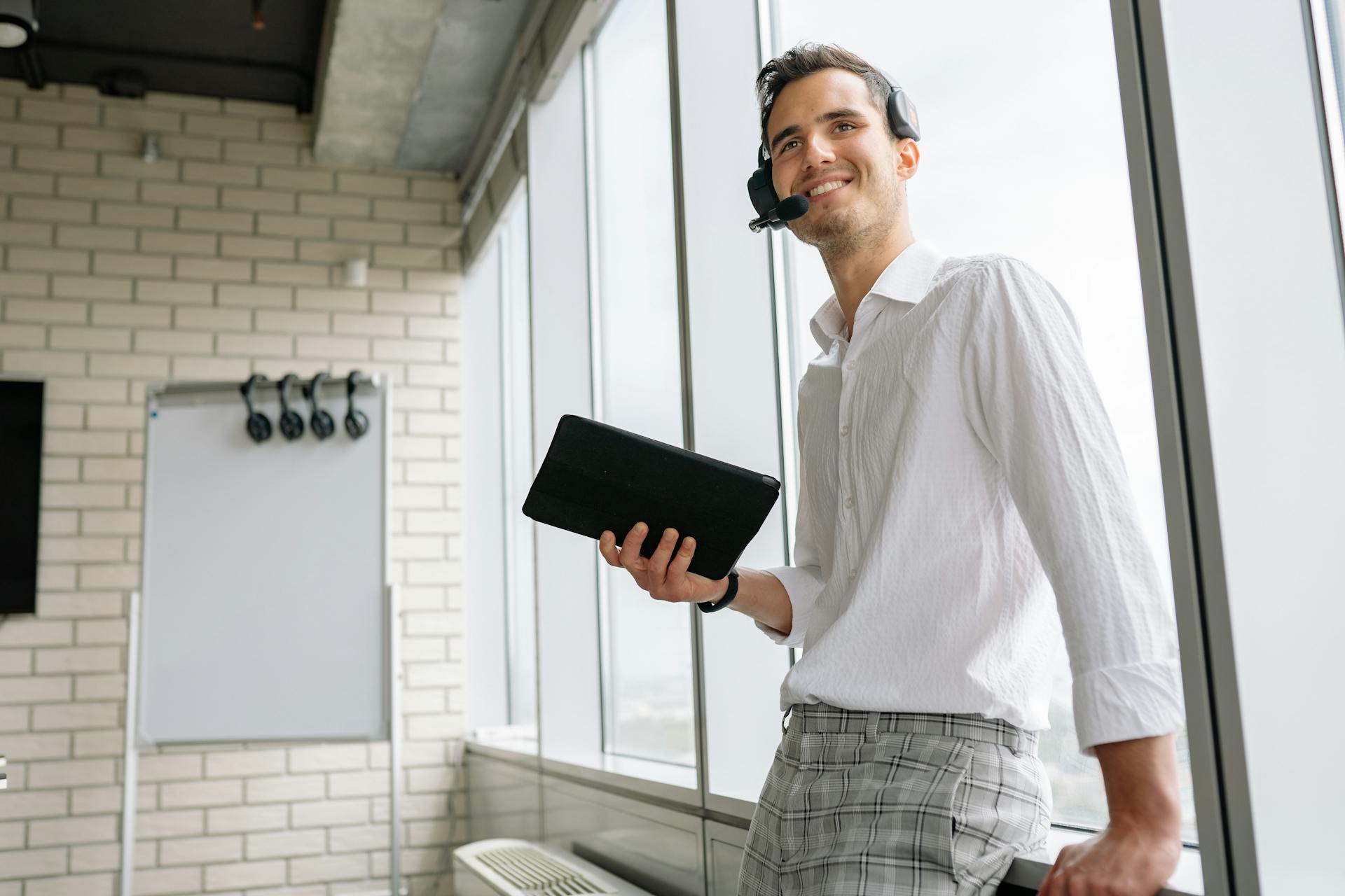 Smiling businessman with a headset and tablet, standing in a modern office environment.