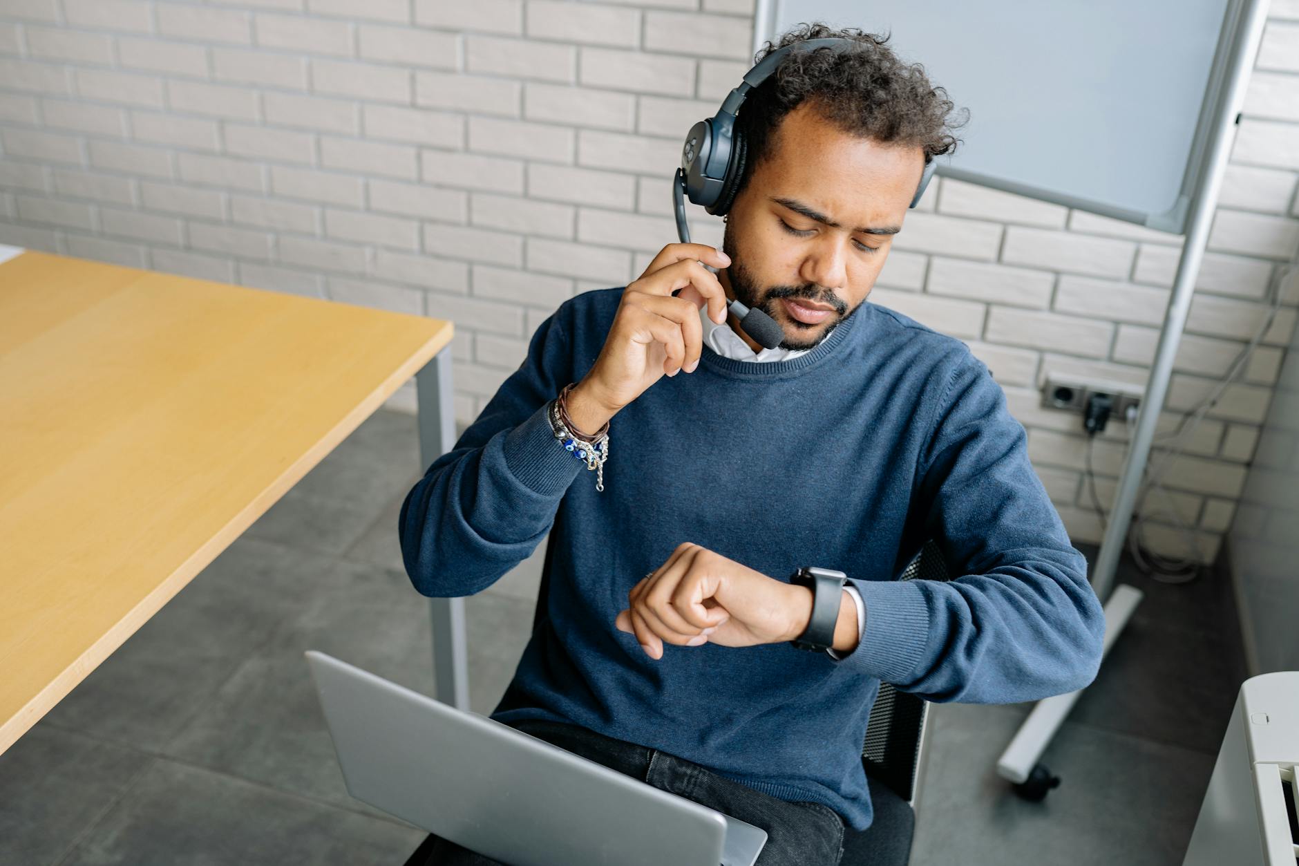 A focused call center agent wearing a headset checks the time on his wristwatch in a modern office setting.