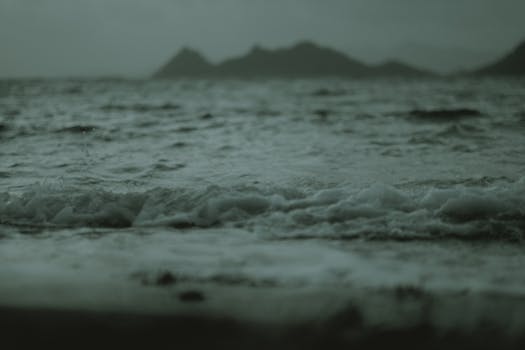 Dramatic ocean waves crashing against the shore at twilight, with distant mountains in the background.