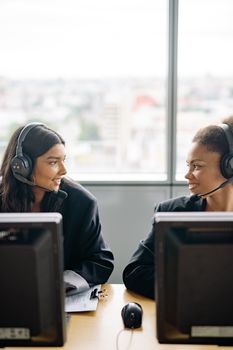 Two colleagues in a call center smiling while working with headsets and computers.