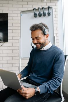 A man in a blue sweater sits in an office, working on a laptop with a headset, smiling.