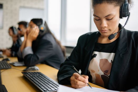 Focused call center employee using headset and writing notes at her desk, working efficiently.