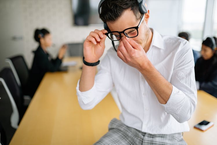 Man In White Long Sleeve Shirt Sitting On A Table And Holding His Nose