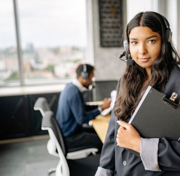 Confident woman wearing a headset and holding a clipboard in a busy office setting.
