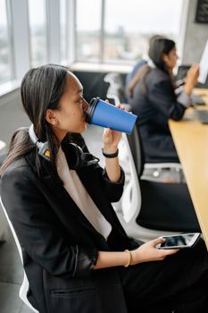 Asian businesswoman in a modern office drinking coffee during a break from customer support work.