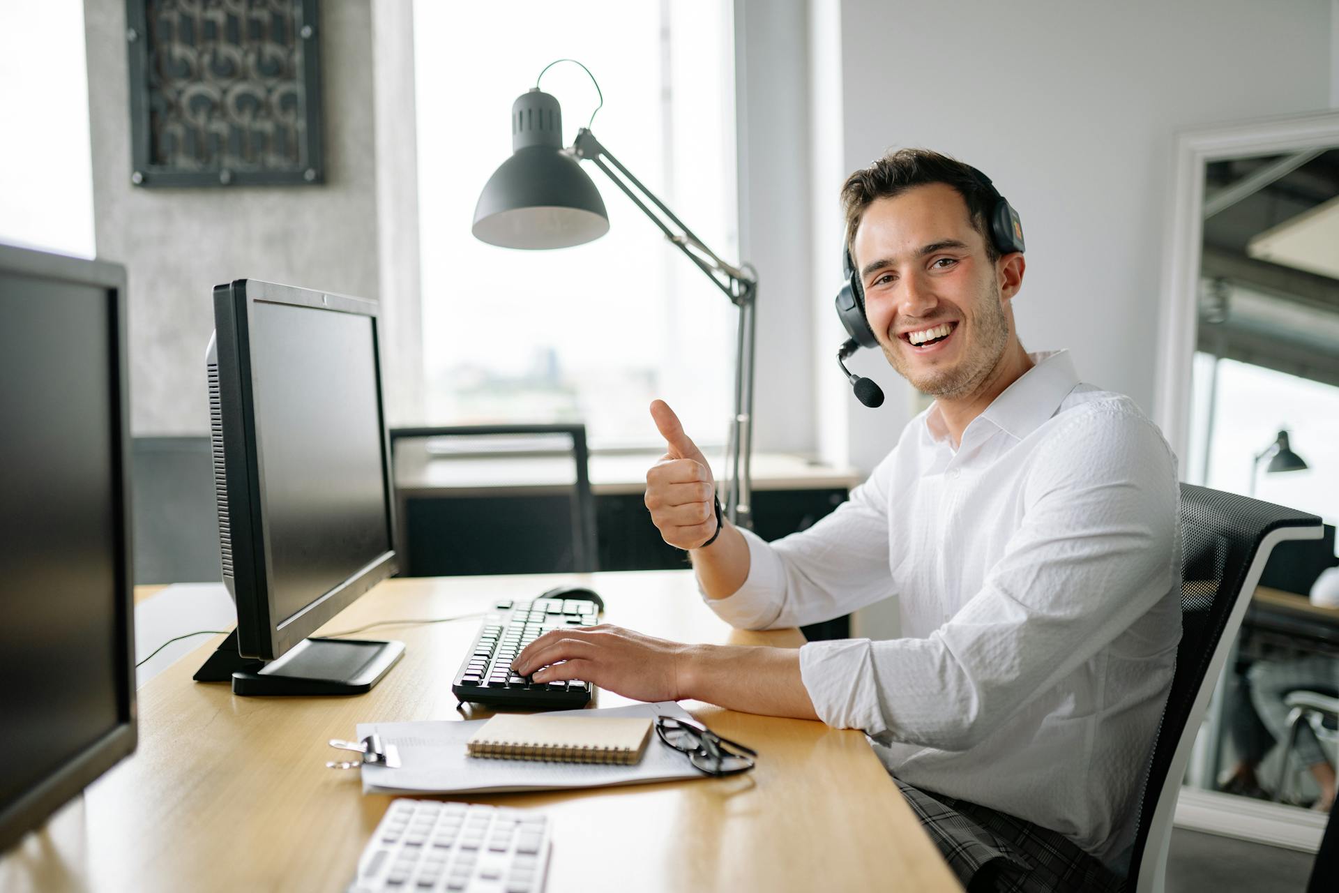 Happy man in office wearing headset, giving a thumbs up while typing at a computer.