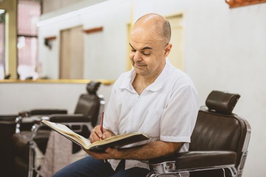 Adult man writing in a notebook while sitting on a barber chair in a well-lit barber shop.