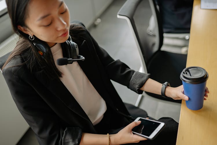 Young Woman Sitting At A Table With A Coffee, Scrolling Through Her Phone And Wearing A Headset 