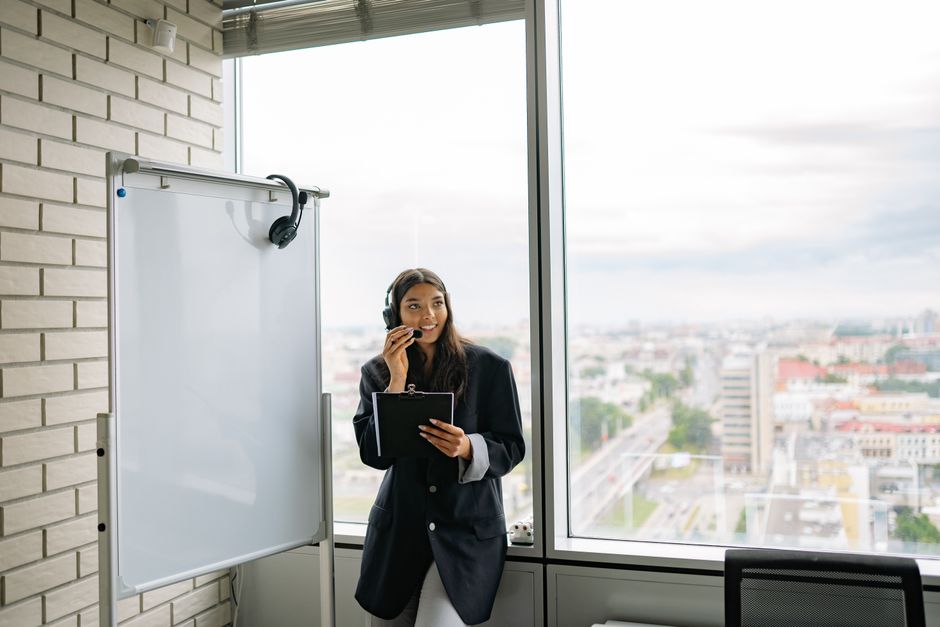 Professional woman wearing headphones standing in a modern office with a city view. Ideal for business themes.
