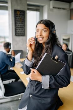 Young woman smiling while using a headset in a modern office, holding a clipboard and working.