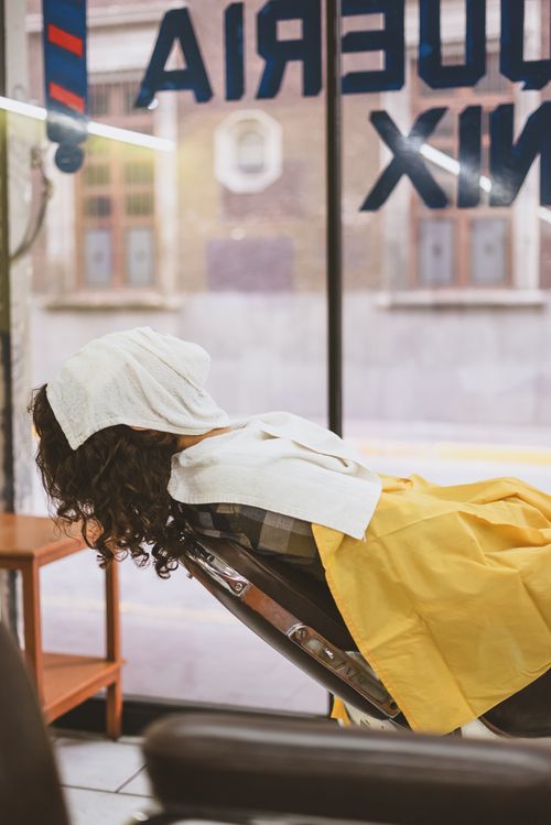 Free A woman with curly hair relaxes in a barbershop chair enjoying self-care with a towel on her head. Stock Photo