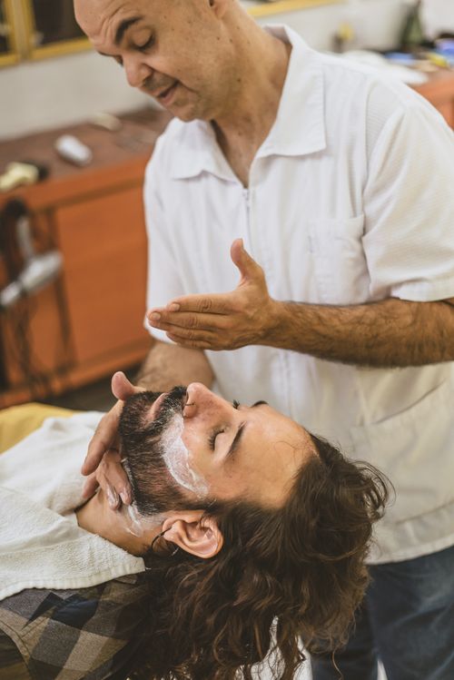 Free Barber applying shaving cream to a male client's beard in a barber shop. Stock Photo