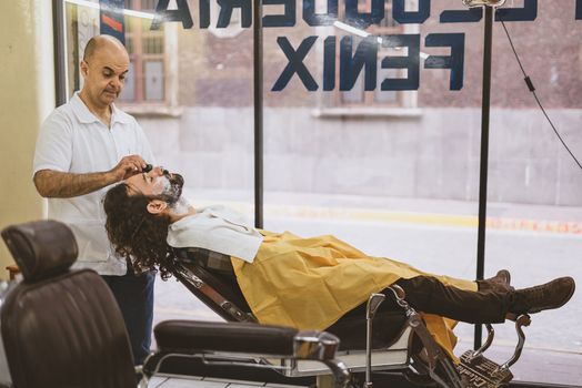 A barber skillfully shaves a client's beard as he relaxes in a barber chair inside a shop.
