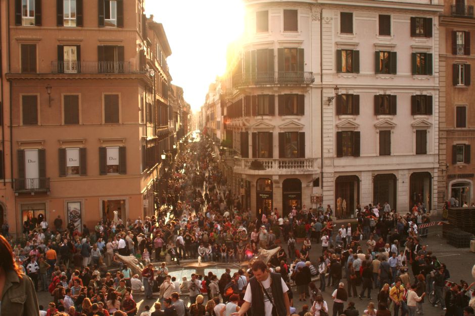 A bustling crowd fills a historic Roman street as the sun sets, creating a vibrant urban atmosphere.