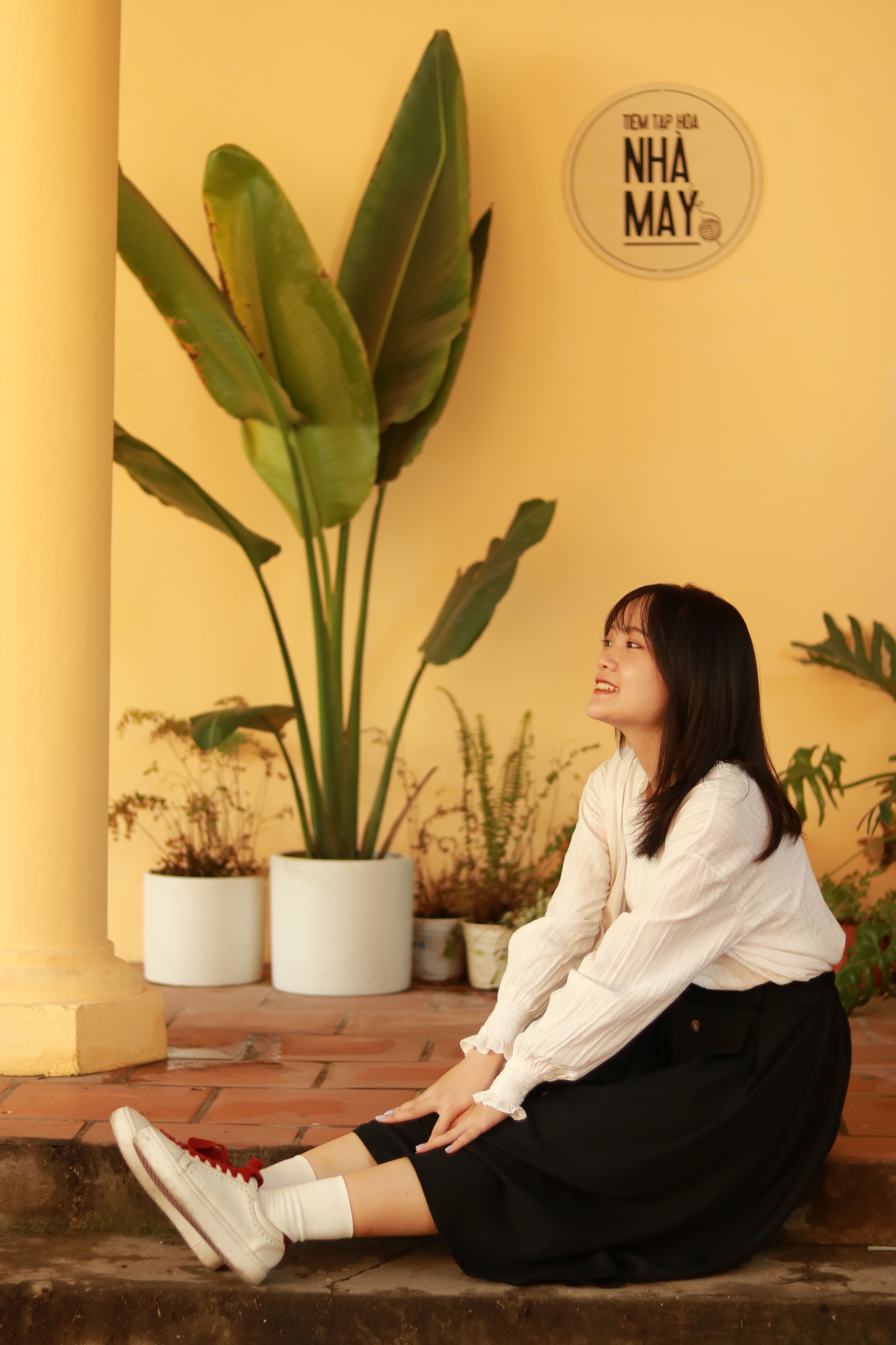 A young woman in casual attire seated beneath indoor plants in a cozy setting.