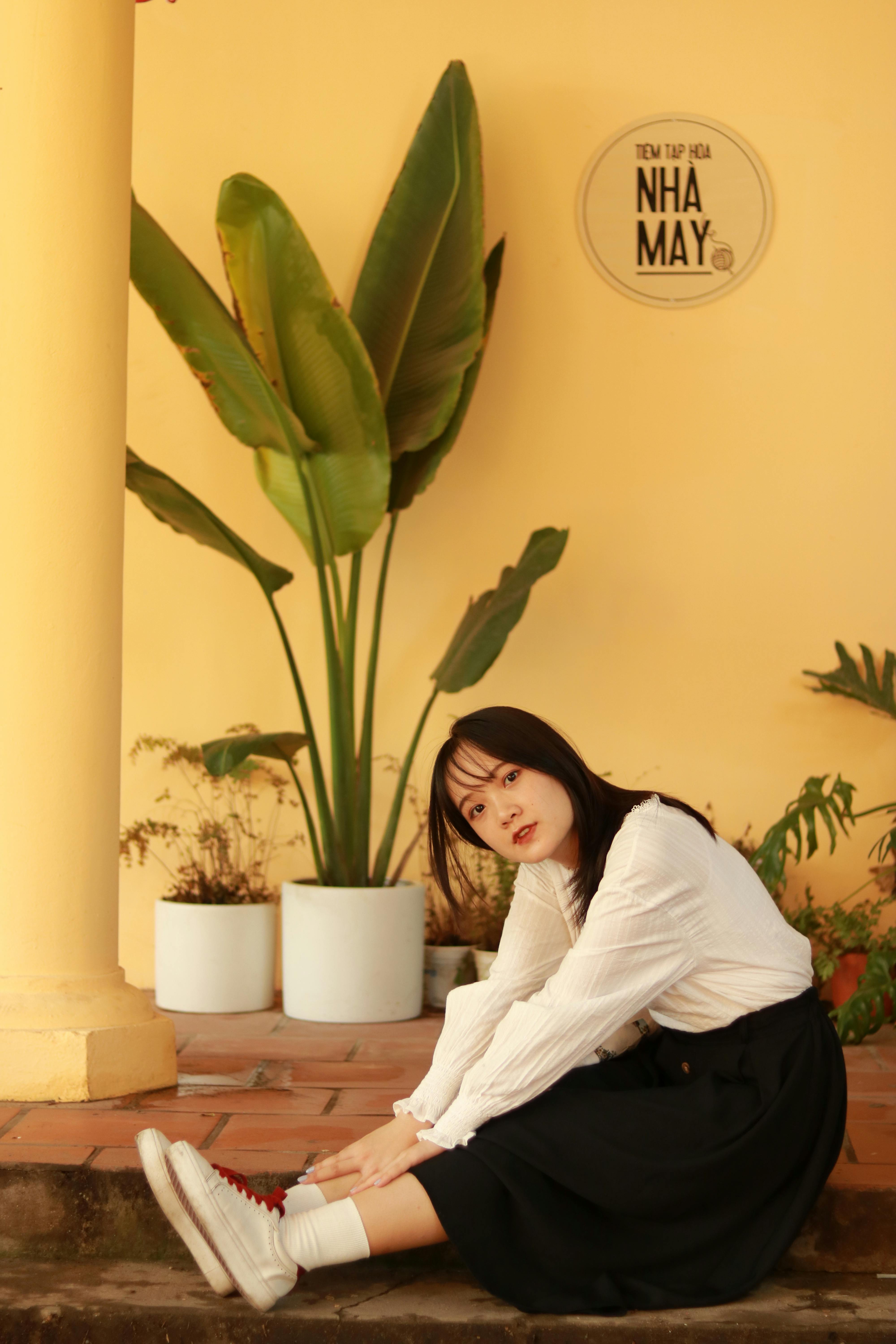 A woman in a stylish outfit sitting indoors with potted plants and yellow walls.
