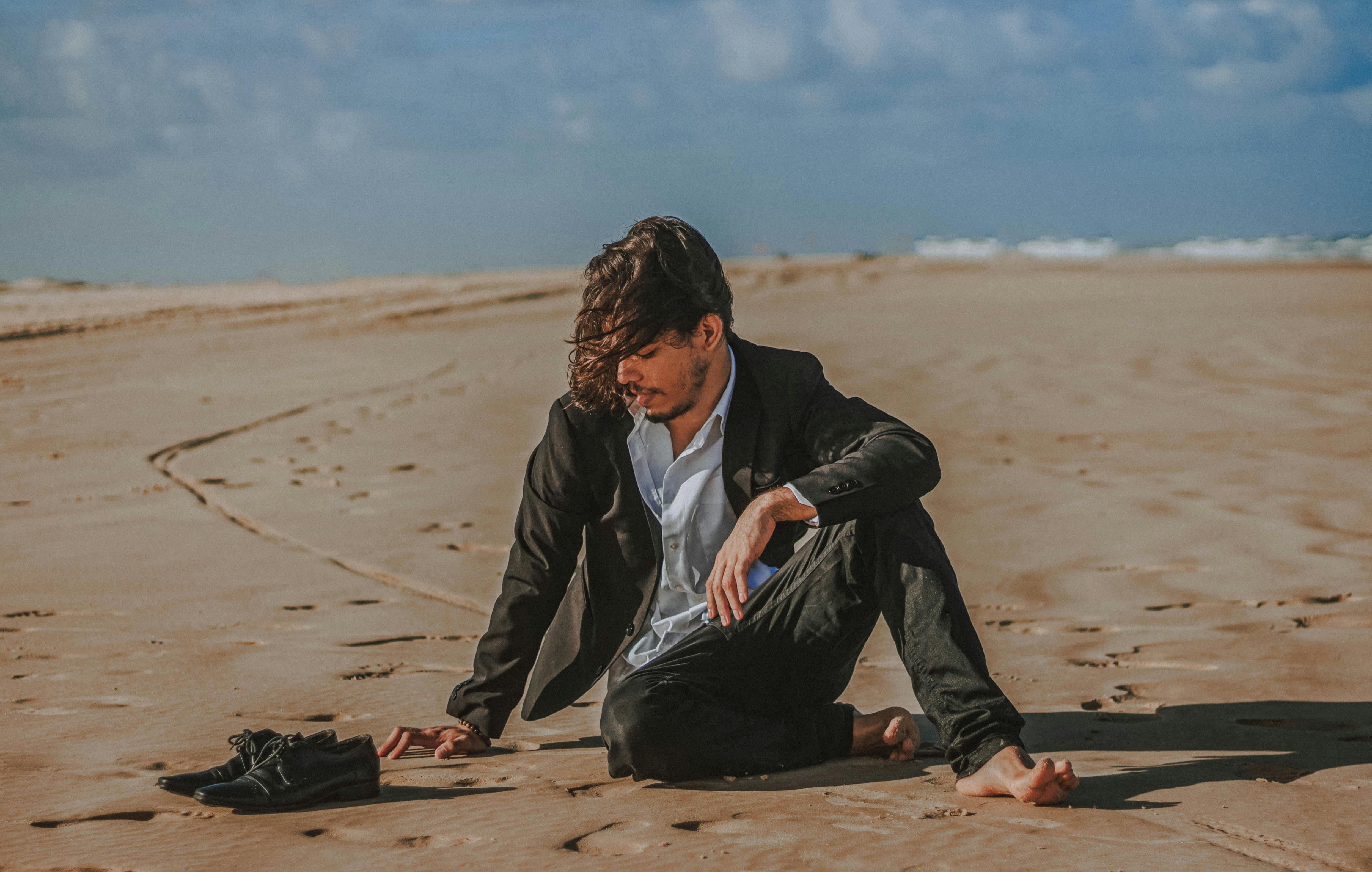 Man in suit sitting barefoot on a sandy beach, contemplating surroundings.