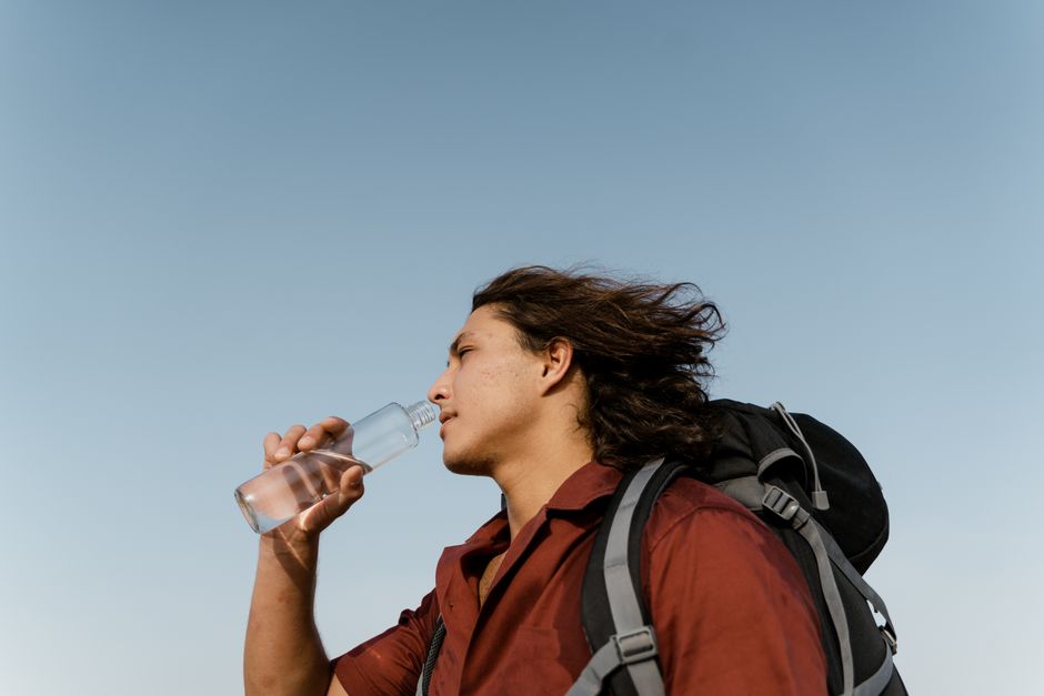 A young man drinking water while hiking with a backpack under the clear sky.