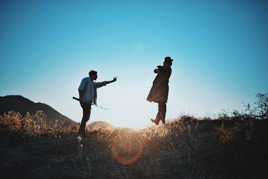Two men engage in a dramatic duel with swords silhouetted against a sunset on a grassy hill.