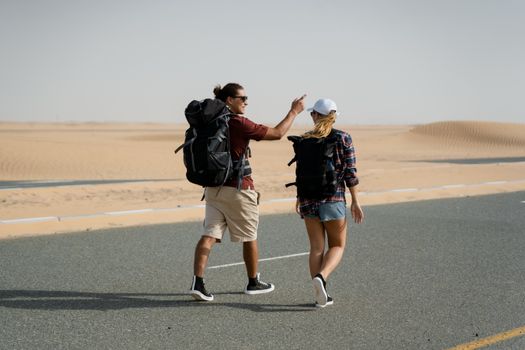 Two travelers with backpacks walk through a desert road in Dubai under a clear sky.